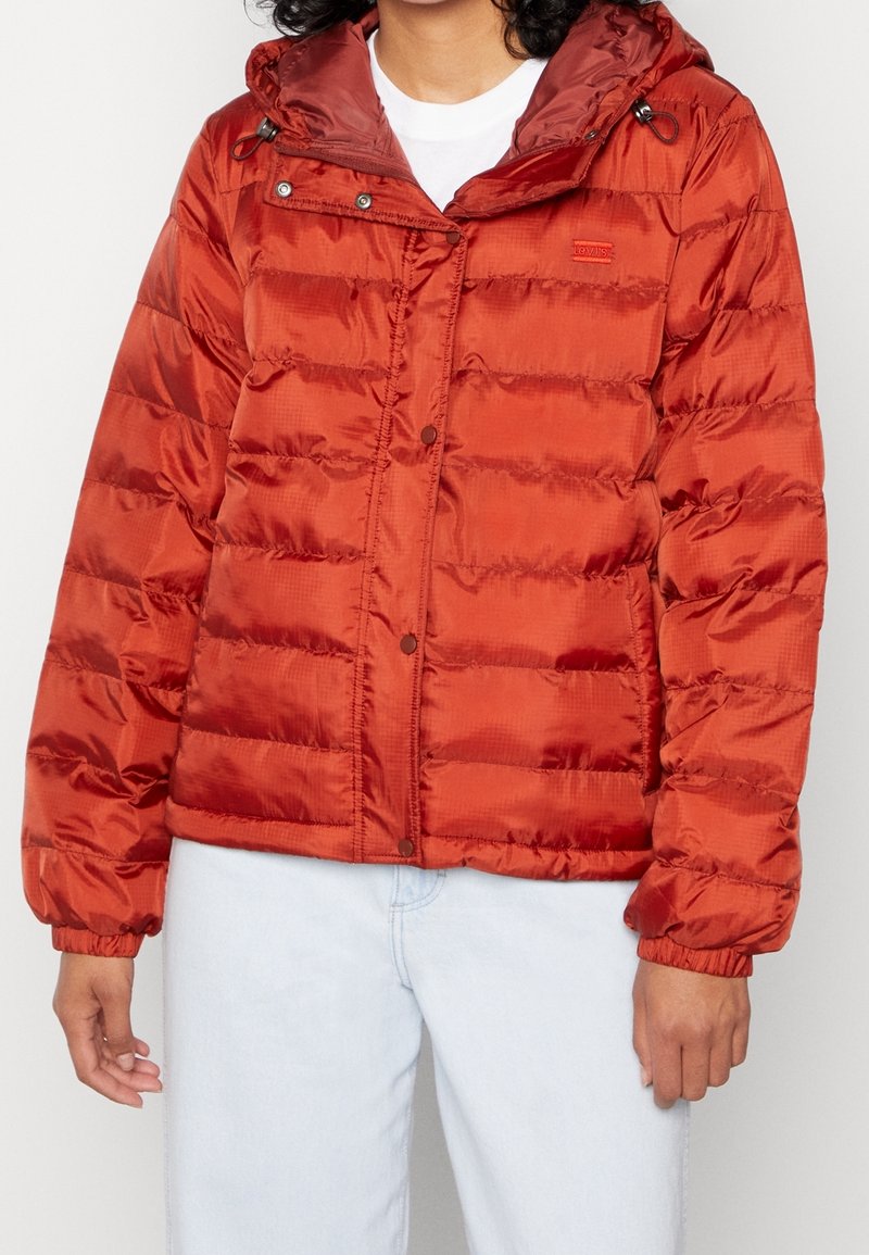 Kvinna som bär en rost-röd vadderad jacka med huva över en vit skjorta och ljusblå jeans.