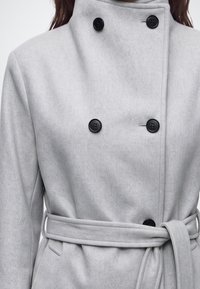 Femme portant un manteau croisé gris clair avec des boutons noirs et une ceinture nouée à la taille.
