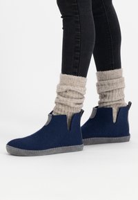 Marineblauwe vilten enkelboots met grijze suède zolen, voorzien van uitsparingen aan de zijkanten en zichtbare stikdetails. Draag met crèmekleurige geribde sokken.