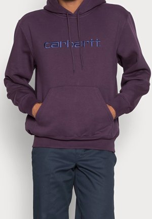 Persona che indossa una felpa con cappuccio Carhartt viola scuro con tasca frontale e pantaloni blu scuro, mani riposte nella tasca della felpa.