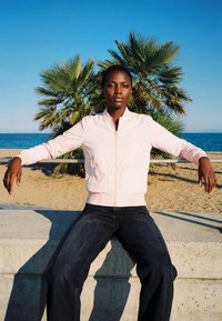 Giovane con le braccia appoggiate su una ringhiera in metallo, vestito con una giacca leggera e pantaloni scuri, seduto su un bordo di cemento vicino alla spiaggia con una palma e l’oceano sullo sfondo.