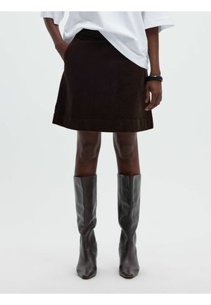 Jupe marron foncé portée avec des bottes noires montantes et un haut blanc oversize, visible de la taille aux pieds sur une personne.