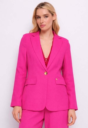 Donna con capelli biondi lunghi indossa un blazer rosa acceso e pantaloni abbinati su una maglia di pizzo rossa, posando contro uno sfondo chiaro.