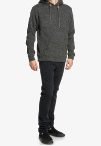 Grauer gestrickter Kapuzenpullover mit einer Kängurutasche, verstellbaren Zugbändern, kombiniert mit dunklen Hosen und schwarzen Slippern.