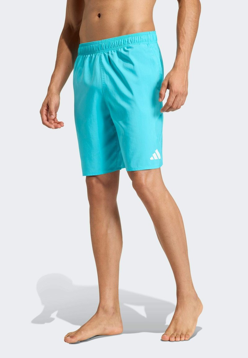 Man draagt turquoise korte broek tot op de knieën met een wit logo op het linkerbeen, staat blootsvoets tegen een effen achtergrond.