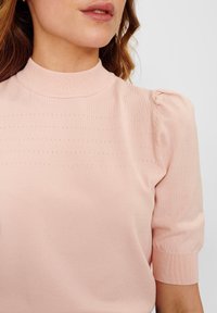 Světle růžový pletený top s vysokým, žebrovaným výstřihem a krátkými, vypouklými rukávy s jemným horizontálním detailem. Hladká textura.