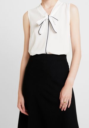 Witte mouwloze blouse met knoopdetail bij de kraag, zwarte bies, gecombineerd met een zwarte aansluitende rok. Gladde stoftextuur.
