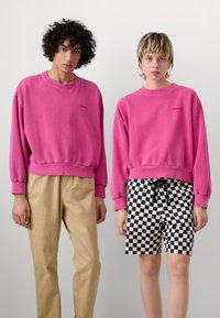 Två modeller bär överdimensionerade rosa sweatshirts med ribbade ärmslut. En matchar det med beiga byxor, medan den andra har på sig svart- och vitrutiga shorts.