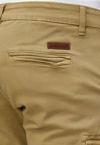 Khaki nadrág, sima textúrával, hátul cipzáras zsebbel és dombornyomott felirattal ellátott barna bőr márkajelzéssel.