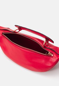 Sac à main en cuir rouge avec une ouverture zippée, quincaillerie dorée et une poignée supérieure. Texture lisse avec un accent noir le long de la poignée.