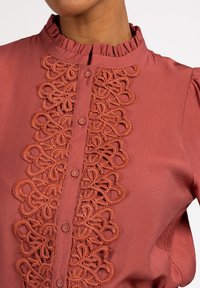 Rustfarbene Bluse mit einer Rüschengenau und aufwendiger Stickerei in der Mitte. Verfügt über vordere Knöpfe und eine weiche Textur.