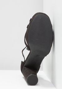 Chaussure à talon haut en daim noir avec bout rond et brides autour de la cheville, vue de la semelle sur fond blanc en angle.