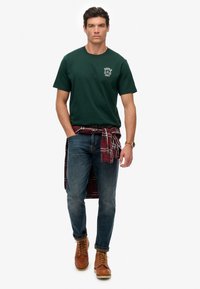 Donkergroen katoenen T-shirt met wit logo, gecombineerd met een blauwe spijkerbroek en bruine laarzen. Een rood en zwart geruit shirt is om de taille gebonden.