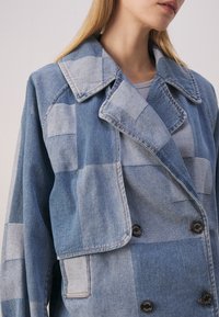 Denim jacket featuring a patchwork design in various shades of blue, with a notched collar and double-breasted button closure.