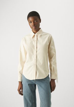 Crème-kleurig langarm shirt met een knoopsluiting aan de voorkant, voorzien van decoratieve stiksels en een gebogen zoom, gecombineerd met lichtblauwe spijkerbroek.