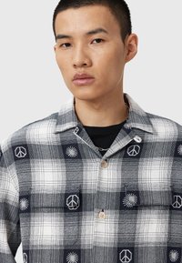 Chemise à carreaux avec des motifs en noir et blanc, boutons en bois et signes de paix et motifs de soleil brodés sur la poitrine.