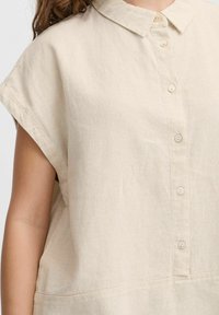 Blusa di lino beige con collo a camicia, maniche corte e chiusura con bottoni sul fronte; presenta un tessuto strutturato e un design a vestibilità rilassata.