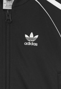 Mustat vetoketjulliset takit, jotka on valmistettu merestä kerätystä muovista. Ominaisuuksiin kuuluu valkoinen Adidas-logo, kontrastivärit olkaimissa ja teksturoitu kangas.