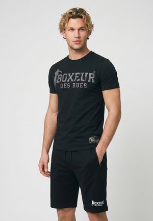 Młody mężczyzna z falującymi blond włosami, ubrany w czarny t-shirt Boxeur des Rues i pasujące czarne spodenki z rękami w kieszeniach.