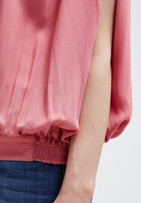 Różowa bluzka bez rękawów z elastycznym dołem noszona przez osobę w niebieskich dżinsach, ukazująca odkryte ramię i detale rozcięcia z boku bluzki.