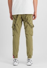 Olive zelené cargo nohavice s viacerými bočnými vreckami, elastickými manžetami a jedinečným detailom pásu na zadnej strane. Stylizované s bielymi teniskami.