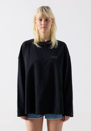 Blonde Frau trägt ein übergroßes schwarzes Langarmshirt mit dem Aufdruck "American Vintage" und blaue Jeansshorts vor weißem Hintergrund.