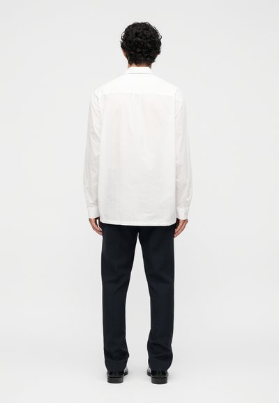 Άνδρας με σγουρά μαλλιά που φοράει λευκό πουκάμισο με μακριά μανίκια, μαύρο παντελόνι και μαύρα παπούτσια, στεκόμενος με την πλάτη σε απλό λευκό φόντο.