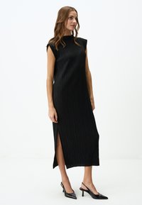 Schwarzes, ärmelloses Midikleid mit gefalteter Textur und seitlichem Schlitz. Verfügt über einen hohen Kragen und strukturierte Schultern. Getragen mit spitzen Slingback-Highheels.