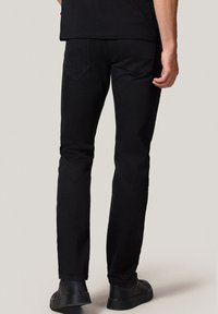 Mężczyzna w czarnych dopasowanych dżinsach i czarnych sneakersach stojący plecami do gładkiego jasnego tła.