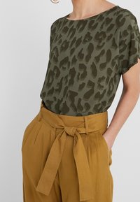 Blusa verde con estampado de leopardo y mangas cortas combinada con pantalones mostaza de cintura alta que cuentan con un cinturón atado y detalles plisados.