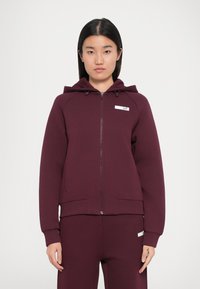 Burgundy zipzáras kapucnis pulóver, elülső zsebekkel és egy kis márkajelzéssel a mellkasán. Puha, strukturált anyag.