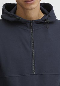Navy kapucnis pulóver, texturált anyagból készült, cipzáros záródással, állítható húzózsinórokkal és magas galléros dizájnnal.