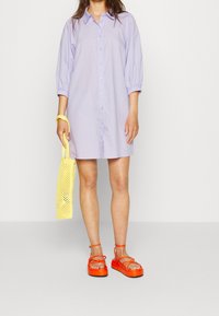 Světle fialové šaty s rukávy do puffu, doplněné jasně oranžovými sandály na řemínky a žlutou síťovanou kabelkou.