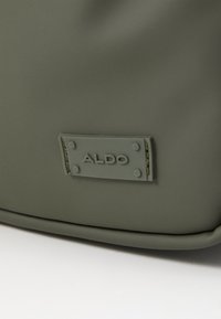 Bolso verde oliva hecho de material suave, con una etiqueta rectangular gris que presenta el texto en relieve "ALDO" y cuatro pequeñas remaches.