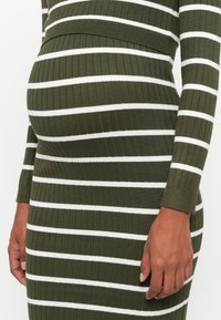 Abito a maglia a costine di colore verde oliva con strisce orizzontali bianche. Il design aderente esalta la forma, con maniche lunghe e scollo a girocollo.