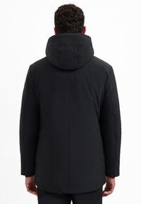 Man met krullend haar in een zwarte winterjas met capuchon, gezien van achteren tegen een eenvoudige witte achtergrond.