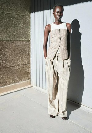 Femme portant un gilet à carreaux beige et un pantalon large, debout contre des murs texturés et lisses sous une lumière vive, les mains dans les poches.