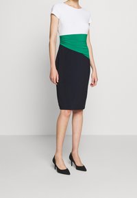 Tajtsittande klänning med vitt överdel, grön midja och mörk marinblå kjol. Tillverkad av stretchig tyg, har en slank silhuett och korta ärmar.