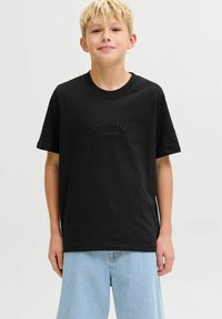 Crna pamučna majica s kratkim rukavima, s reljefnim logotipom "ORIGINALS Studio" na prednjoj strani. Uparena s svijetloplavim trapericama.