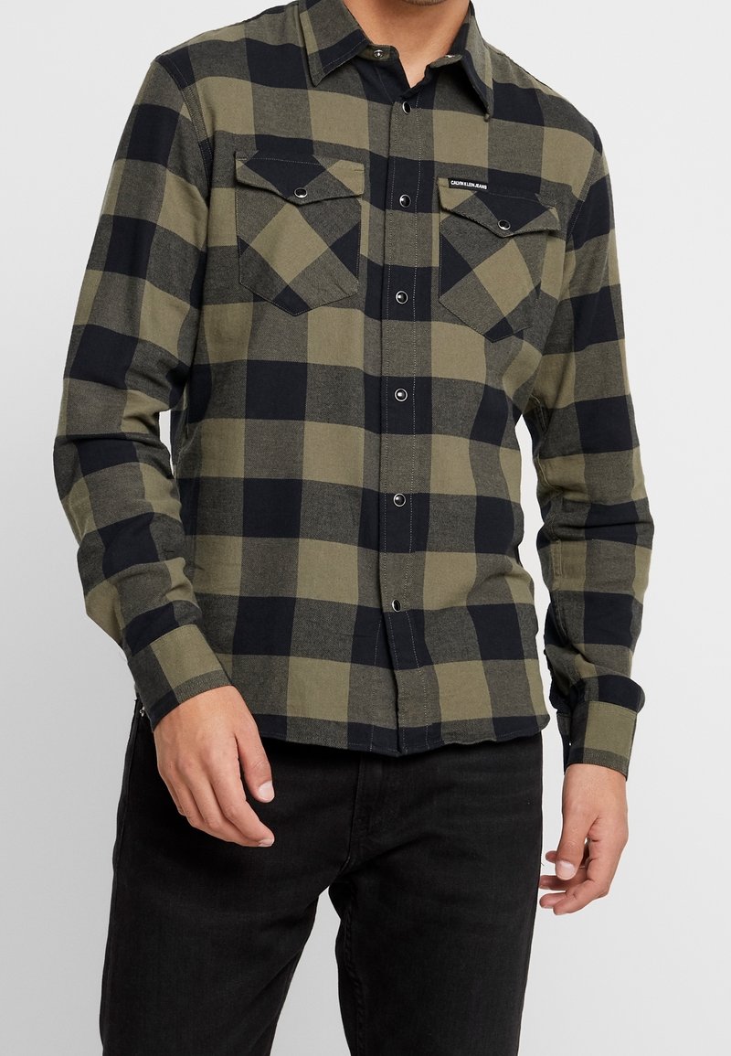Camisa de franela a cuadros verde y negra con mangas largas, cierre de botones en el frente, dos bolsillos en el pecho y detalles de botones metálicos.