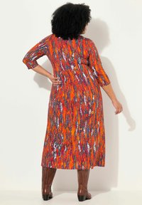 Robe longue et fluide avec un motif vibrant orange et bleu. Dotée de manches trois-quarts et d'une taille cintrée. Portée avec des bottes marron hautes.