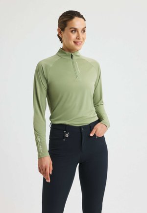 Green long-sleeve top with a half-zip collar and raglan sleeves; paired with navy fitted pants featuring decorative stitching and pocket details.