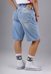 Jeansshorts in Hellblau, mit hoher Taille, fünf Taschen und einem Lederpatch auf der Rückseite. Kombiniert mit weißen High-Top-Sneakern und Socken.