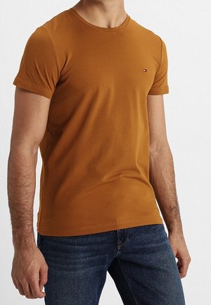 Mężczyzna w dopasowanym musztardowym t-shircie z krótkim rękawem i małym logo na piersi oraz niebieskich dżinsach, stoi na jednolitym tle.