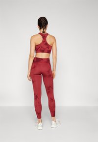 Kobieta z warkoczami, ubrana w sportowy biustonosz w czerwonej kolorystyce tie-dye i dopasowane legginsy, stojąca tyłem na jednolitym białym tle.