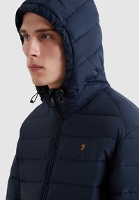 Marinefarbener, gepolsterter Kapuzenjacke mit horizontalen Nähten; verfügt über einen Reißverschluss und eine verstellbare Kapuze. Kleines oranges Logo auf der linken Brust.