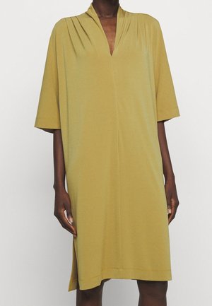 Løs pasform olivengrøn knælængde kjole med albuelange ærmer og en dyb V-hals, båret af en mørkhudet person.