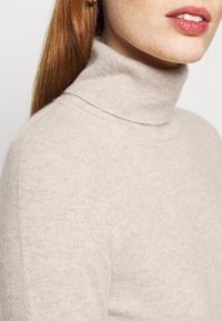 Gros plan d'une personne portant un pull à col roulé beige avec de longs cheveux rouges tirés en arrière et des lèvres roses brillantes visibles.