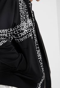 Czarna bluza z kapturem na zamek, z białymi grafikami na rękawach, wykonana z gładkiego, miękkiego materiału o luźnym kroju i z kieszeniami.