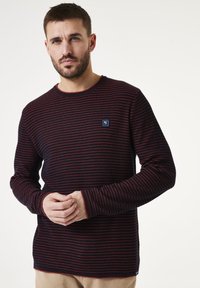 Suéter a rayas en azul marino con acentos rojos, hecho de un tejido de punto texturizado. Presenta un cuello redondo y un pequeño parche con logo en el pecho.